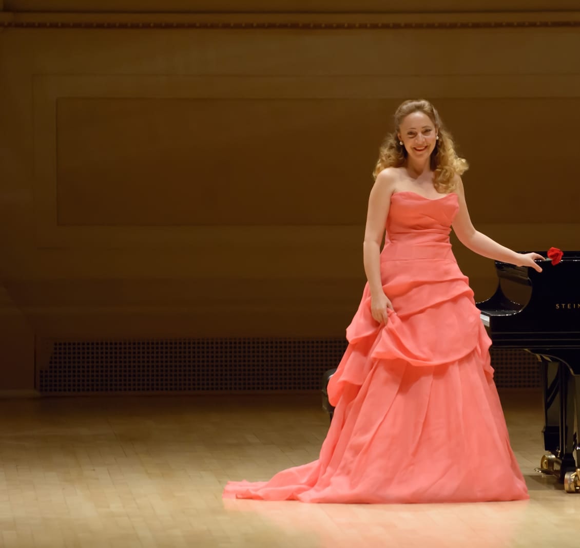 Katya Grineva in a coral gown beside the piano, holding a single red rose
