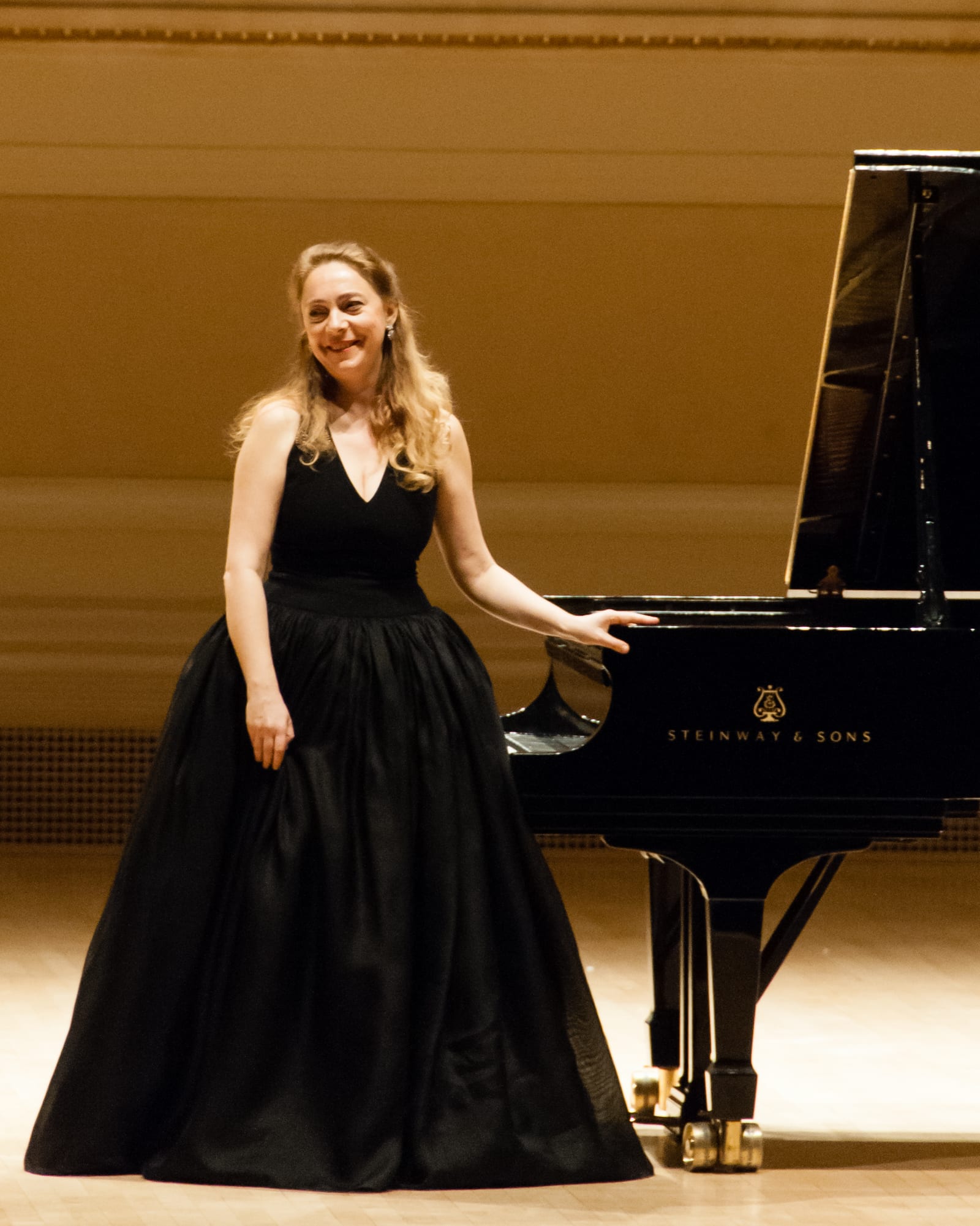Katya Grineva in a black ball gown standing beside a Steinway grand, the gilt hall warm behind her