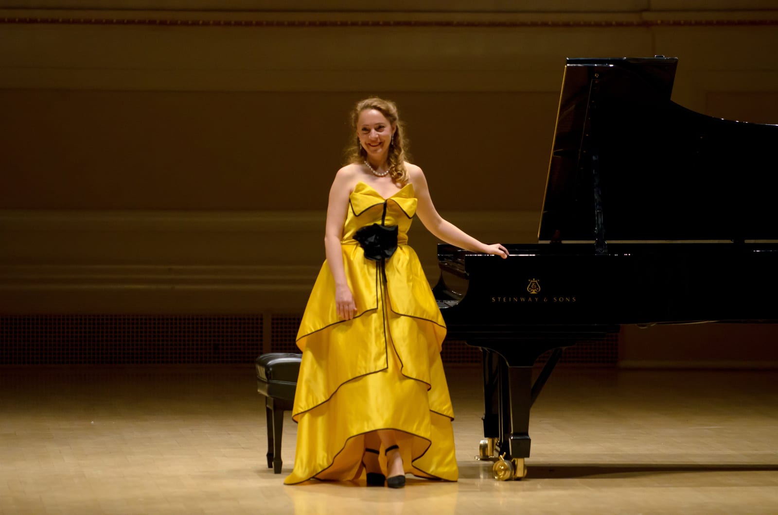 Katya Grineva in a yellow tiered gown beside the Steinway on the Carnegie Hall stage