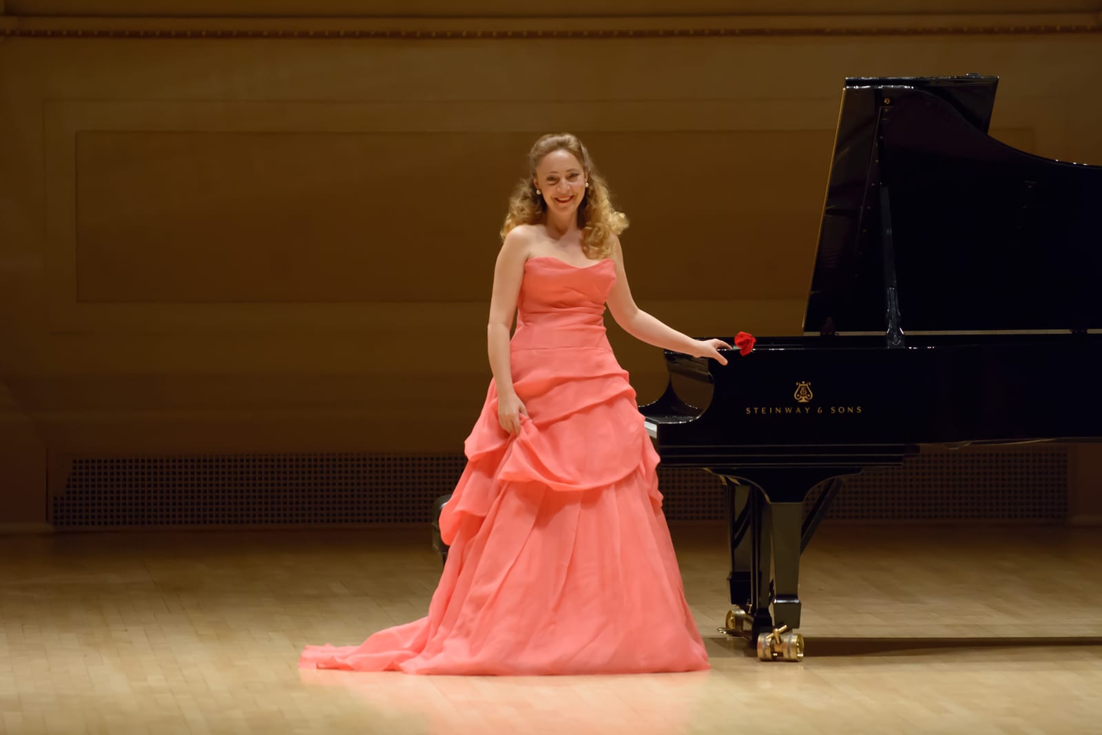 Katya Grineva in a coral gown beside the piano, holding a single red rose