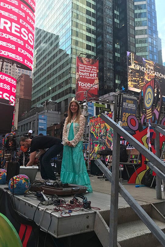 Katya Grineva on an outdoor platform in Times Square while a crew sets up a keyboard, billboards rising behind