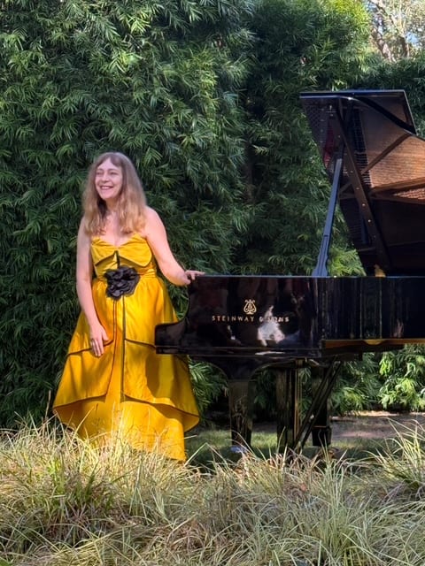 Katya Grineva in a yellow gown standing at a Steinway among the hedges of a California garden