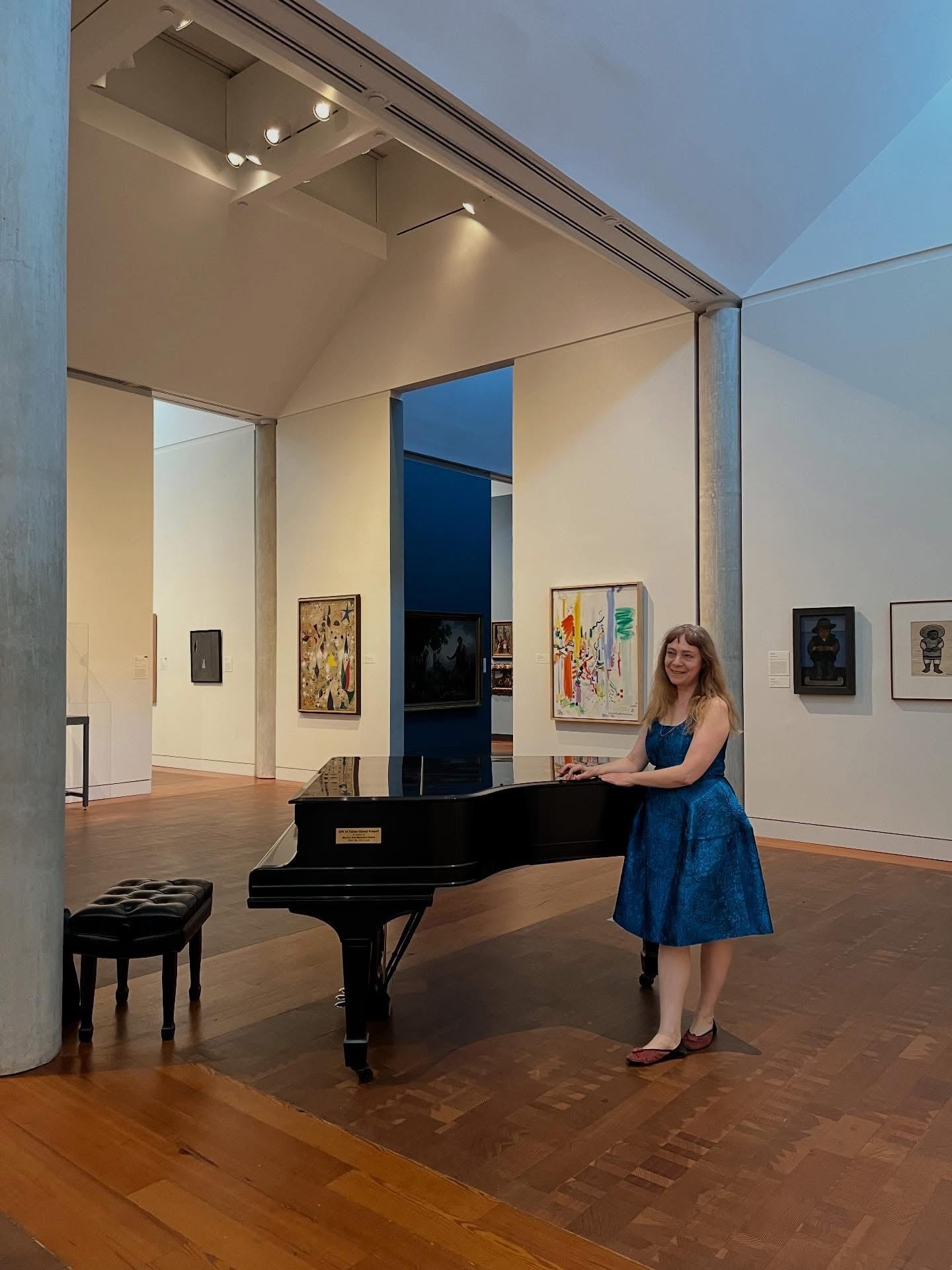 Katya Grineva in a blue dress at a grand piano in a gallery hall hung with paintings