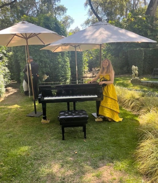 Katya Grineva in a yellow gown beside a piano under white garden umbrellas