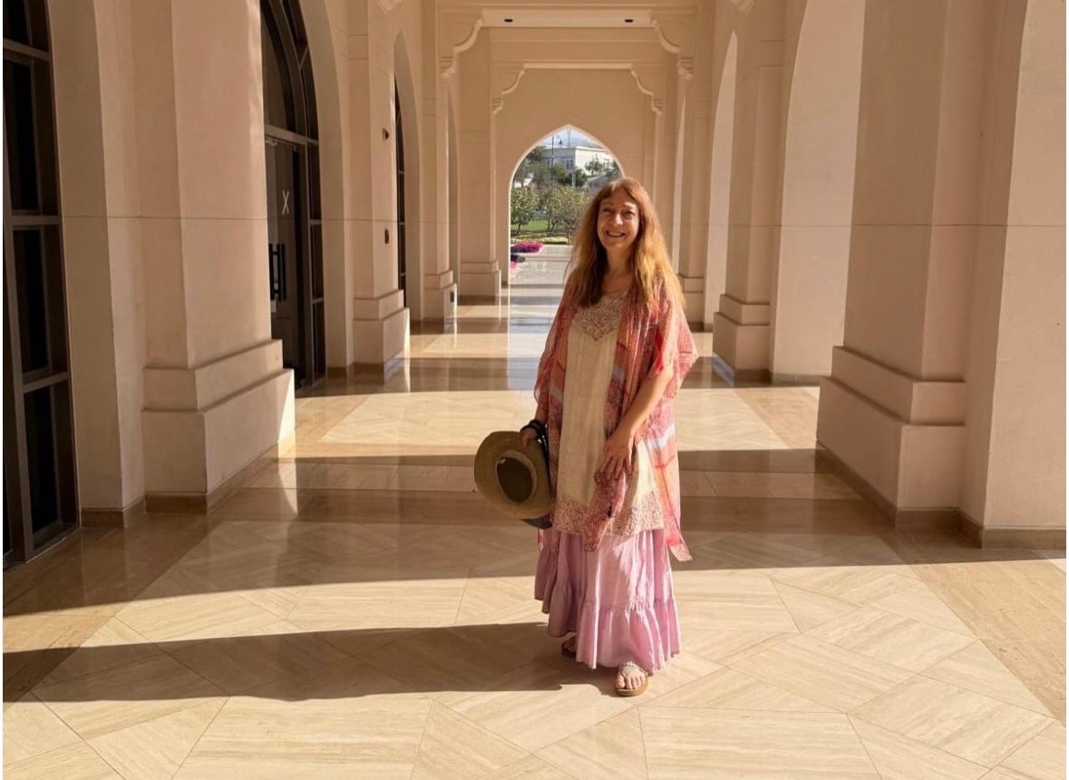 Katya Grineva in a long arched colonnade in India, holding a sun hat