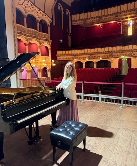 Katya Grineva beside the piano on the stage of the Royal Opera House, red and gold balconies behind her